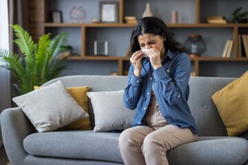 Sick young Indian woman sits on the couch at home and wipes her nose with a tissue from a runny nose