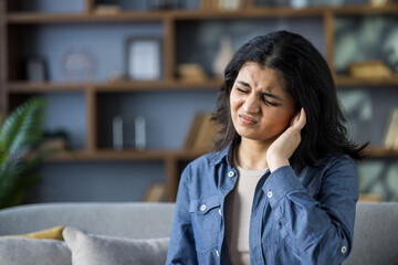 Young Indian woman sitting on sofa at home and holding her ear with her hand, feeling severe pain...