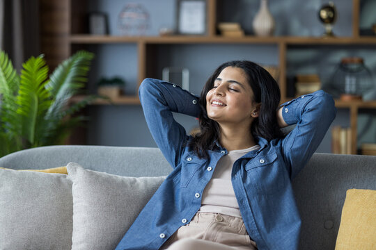 Relaxed young Indian girl sitting on the couch at home, hands behind her head, eyes closed and resting