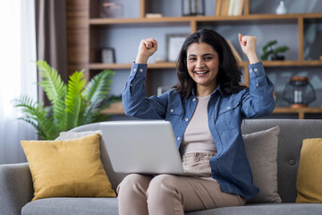 Smiling young Indian girl sitting on the couch at home. holding a laptop and rejoicing in success...