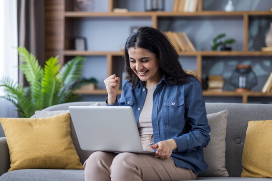 Happy Indian young woman sitting on the sofa at home, holding a laptop on her lap and rejoicing in success, showing a victory gesture