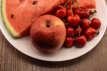a watermelon slice, a red apple, and a cluster red cherries, presented on a white plate with a wooden background. Ideal for themes of wholesome eating, fresh produce, and natural snacks