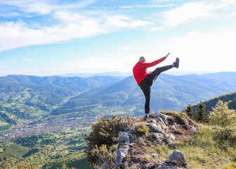 Man in red jacket performing high kick on mountain peak with panoramic view — symbol of strength,...