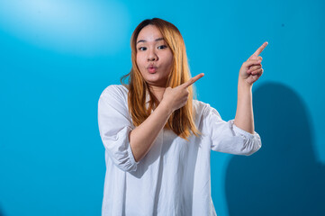 Asian woman with long straight hair wearing casual white shirt smiles brightly while pointing sideways with both hands against vibrant blue background, creating a cheerful and dynamic pose