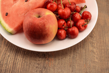a watermelon slice, a red apple, and a cluster red cherries, presented on a white plate with a wooden background. Ideal for themes of wholesome eating, fresh produce, and natural snacks