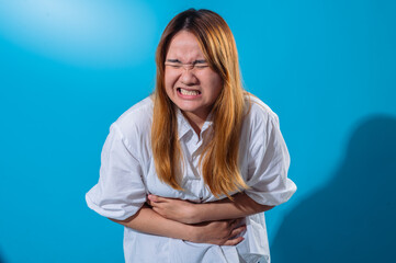 Asian woman in white shirt holding her stomach in pain with a tense facial expression, eyes closed and teeth clenched, expressing abdominal discomfort in front of a solid blue background