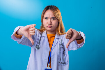 Asian female doctor wearing white coat and orange shirt making double thumbs down gesture with disapproving facial expression, standing against bright blue studio background