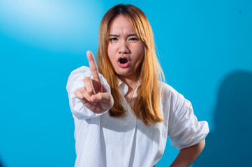 Asian woman in a white shirt pointing her index finger forward with a stern expression, as if warning or scolding, against a plain bright blue background