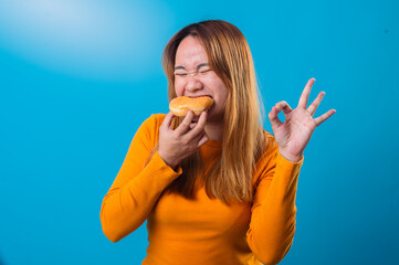 Asian woman in an orange shirt biting a donut and showing an OK hand sign with a satisfied expression, standing against a bright blue background