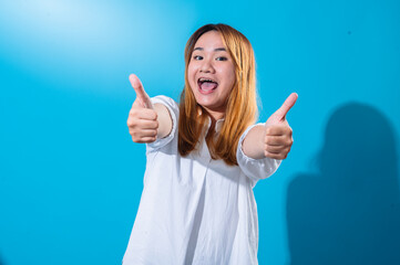Asian woman with long straight hair in white shirt smiles and enthusiastically gives double thumbs up with extended arms, posing against a solid bright blue background in a cheerful studio setting