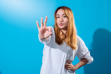 Asian woman with long straight hair in a white shirt smiles brightly while making an OK hand...