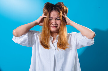Asian woman in white shirt grabbing her head with both hands and gritting her teeth in frustration or stress, eyes shut tightly, standing against a bright blue background