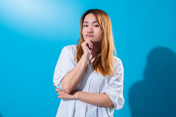Asian woman with long straight hair in white shirt poses with thoughtful expression, resting chin on hand while looking up, standing against bright blue background in studio lighting