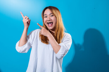 Asian woman with long straight hair wearing casual white shirt smiles brightly while pointing sideways with both hands against vibrant blue background, creating a cheerful and dynamic pose