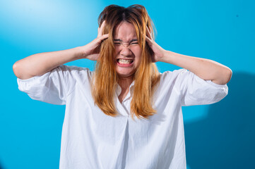 Asian woman in white shirt grabbing her head with both hands and gritting her teeth in frustration or stress, eyes shut tightly, standing against a bright blue background
