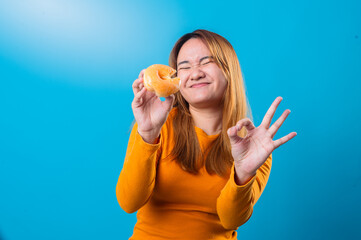 Asian woman in orange long-sleeve shirt holding a bitten glazed donut with a cheeky smile and making an OK hand gesture, posing against a bright blue background