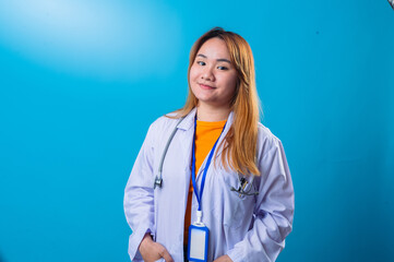 Asian female doctor in white coat with stethoscope around neck, smiling confidently in front of bright blue background, wearing orange shirt and blue lanyard in studio portrait