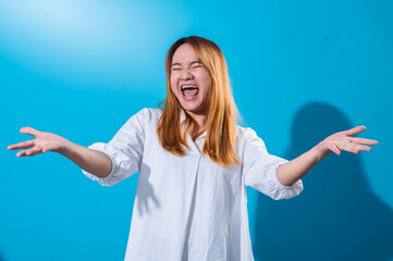 Asian woman with long straight hair wearing white shirt smiles widely while holding both hands open to the sides in welcoming gesture, standing against a bright blue background in studio light