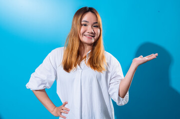 Asian woman with long straight hair in white shirt smiling while holding one hand open to the side as if presenting or showing something, standing confidently against a solid blue studio background