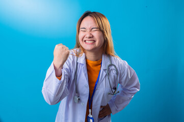 Smiling Asian female doctor celebrating success with clenched fist, eyes closed in joyful expression, wearing white coat, stethoscope, orange shirt, and blue lanyard on blue background