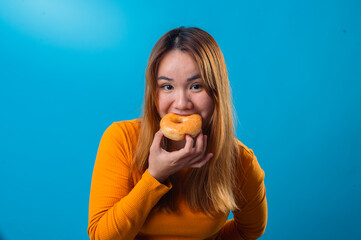 Asian woman in orange sweater bites into a glazed donut with eyes closed and a playful expression, standing against a solid blue background
