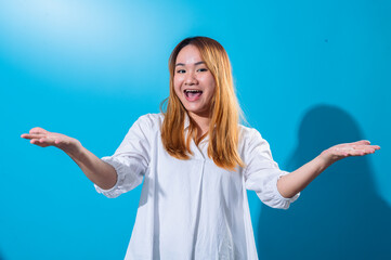 Fototapeta premium Asian woman with long straight hair wearing white shirt smiles widely while holding both hands open to the sides in welcoming gesture, standing against a bright blue background in studio light