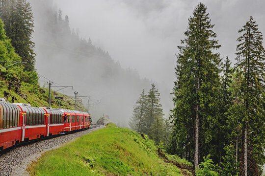 A red train winds along a narrow track amidst a misty mountain landscape