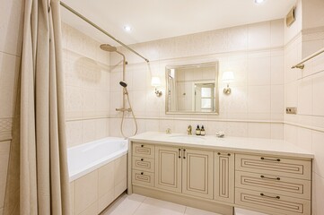 Bright bathroom with beige tile, bathtub with curtain, vanity, and a mirror with sconces
