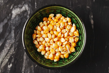 Raw popcorn kernels in bowl. Yellow corn seeds on black marble background. Top view of maize for popping. Natural organic food for homemade snacks. Ready to be turned into crunchy popcorn.