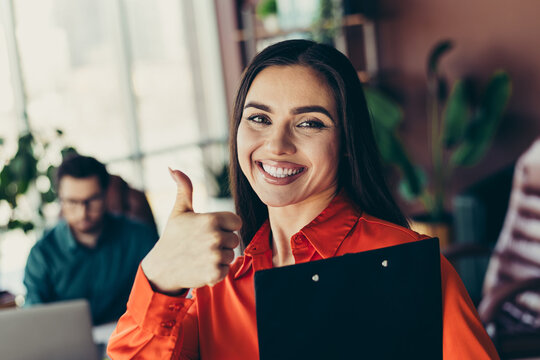 Confident businesswoman smiling in a modern workspace, holding a clipboard and giving a thumbs up as colleagues work nearby.