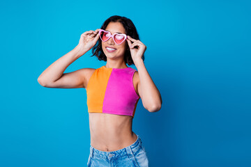 Young woman in summer fashion smiling and posing against a vibrant blue background holding heart-shaped sunglasses