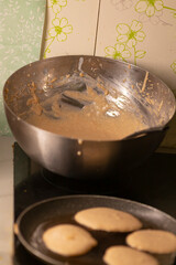 A metal bowl filled with batter alongside a skillet on the stove, which contains freshly cooked circular food items, illustrating the various steps in the food preparation process