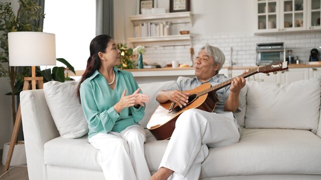 Happy senior man sitting at sofa and playing guitar or music instrument while his wife singing and moving to music. Elder couple spending time together and enjoy doing activity together. Myrmidon.
