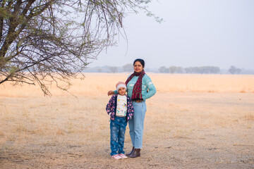 Asian Mother and Daughter wore winter dress sweater jacket cap scarf jeans and boot standing in the background of dry grassland during safari vacation trip. Travel, fashion, parenting concept