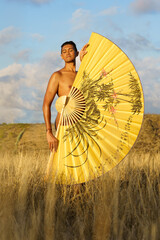 Jeune homme dévêtu rêvant dans la nature, caché derrière un grand éventail jaune