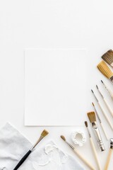 A blank white canvas and a collection of artist's brushes are arranged on a white surface, alongside a piece of gray fabric and a small blob of white paint