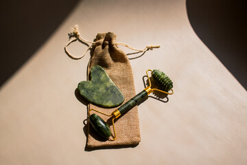 Close up of green quartz gua sha and facial massager on minimalist surface under straight sunlight with hard shadows. Image for cosmetic promotions anti-aging treatments.