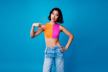 Young stylish woman pointing at herself in vibrant outfit against blue background, showcasing...