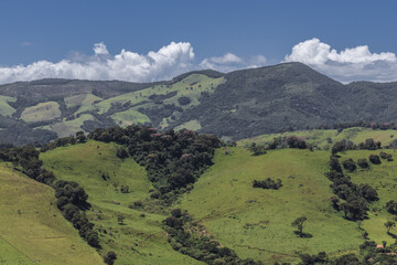 A scenic landscape of rolling green hills and mountains captured in Extrema, Minas Gerais, Brazil. The image shows lush pastureland, forest patches, and dramatic cloud formations against a deep blue