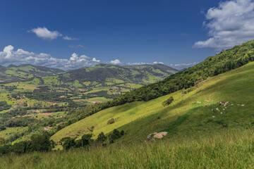 Fototapeta premium A scenic landscape of rolling green hills and mountains captured in Extrema, Minas Gerais, Brazil. The image shows lush pastureland, forest patches, and dramatic cloud formations against a deep blue