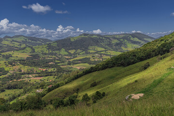 A scenic landscape of rolling green hills and mountains captured in Extrema, Minas Gerais, Brazil. The image shows lush pastureland, forest patches, and dramatic cloud formations against a deep blue