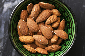 Whole almonds in green bowl. Natural unpeeled almonds on black marble background. Top view healthy organic snack. Nutritious food full of energy. Rustic style with oriental dish aesthetic.