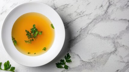 A clear broth soup garnished with fresh parsley leaves served in a white bowl on a marble surface.