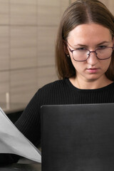 Young woman accountant wearing eyeglasses working from home, reviewing financial documents and using laptop, performing accounting tasks remotely. Vertical photo