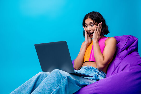 Young woman on colorful beanbag with surprised expression using laptop against bright blue background