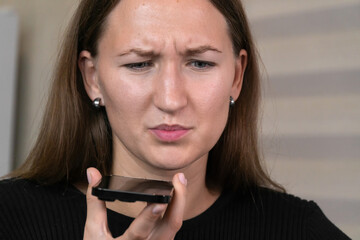 Focused young woman using a smartphone in the kitchen, recording an audio message or making a phone call on speakerphone, enjoying the convenience of modern communication technology