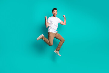 Handsome young man jumps joyfully against a vibrant teal background wearing casual attire and...
