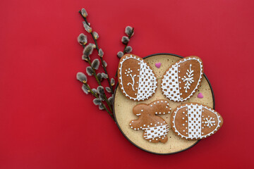 Romantic decorated Easter gingerbread cookies in a shape of egg and bunny with small sugar hearts on a plate, and willow twigs on a red background, copy space for text