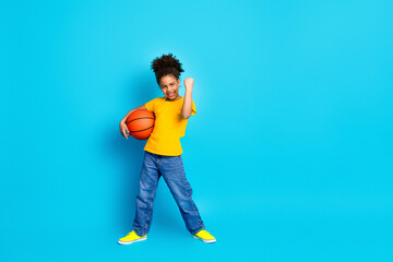 Preteen girl with basketball showing a cheerful gesture on a vibrant blue background, wearing casual yellow and denim clothes