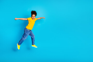 Obraz premium Cheerful young girl jumping with enthusiasm on a vibrant blue background, wearing casual yellow shirt and jeans, expressing energy